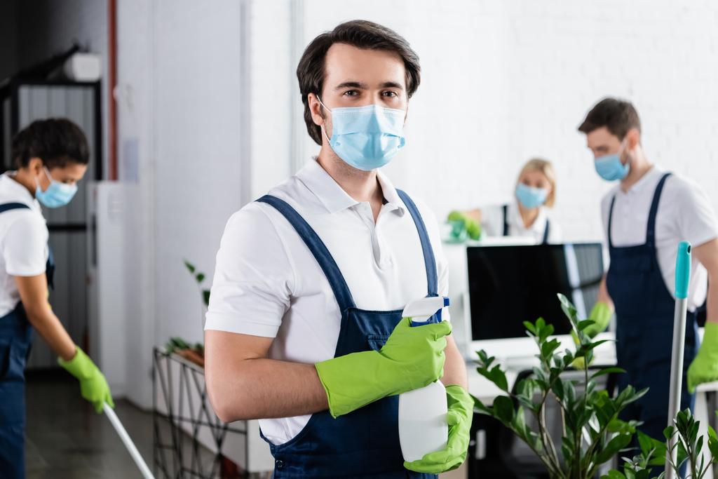 Работник чистящей компании в медицинской маске, держащий моющее средство в офисе  - Фото, изображение