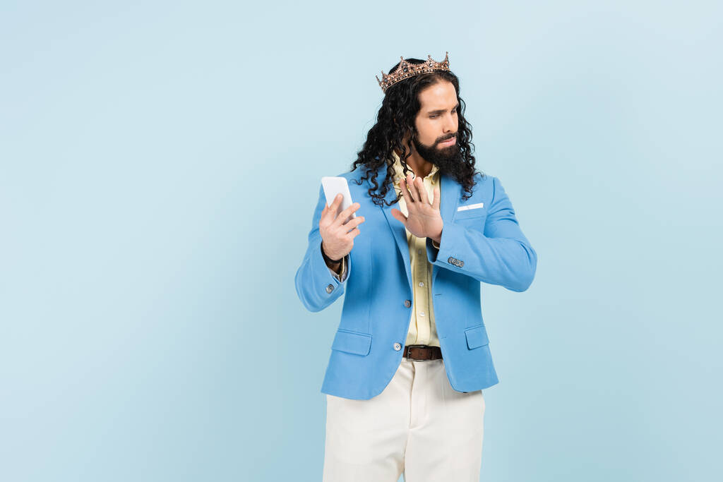 beledigd latino man in jas en kroon tonen stop gebaar terwijl het houden van smartphone geïsoleerd op blauw - Foto, afbeelding