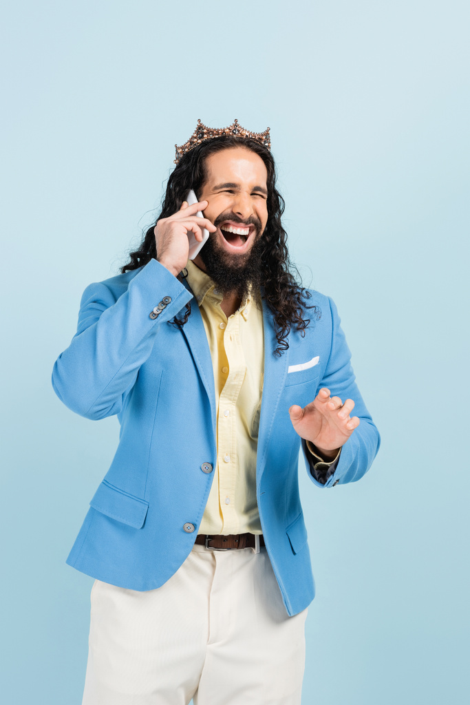 onnellinen latino mies takki ja kruunu puhuu älypuhelimella ja nauraa eristetty sininen - Valokuva, kuva