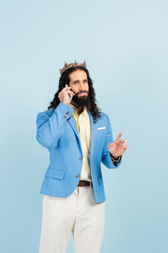 γενειοφόρος ισπανόφωνος με σακάκι και στέμμα μιλάει στο κινητό απομονωμένος στο μπλε - Φωτογραφία, εικόνα