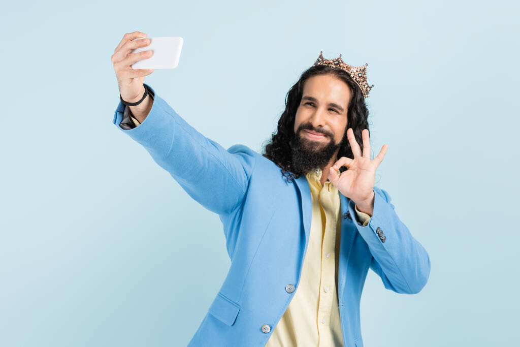 happy hispanic man in jacket and crown showing okay and taking selfie isolated on blue - Photo, Image