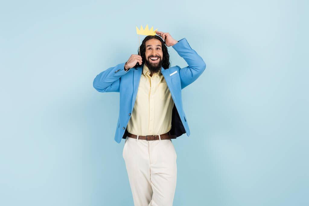 szczęśliwy Latynos człowiek w kurtce trzymając papierową koronę na patyku odizolowany na niebiesko - Zdjęcie, obraz
