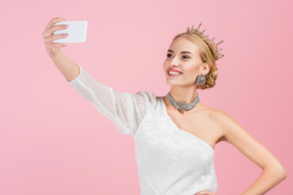 blonde woman in luxury crown taking selfie on smartphone isolated on pink - Photo, Image