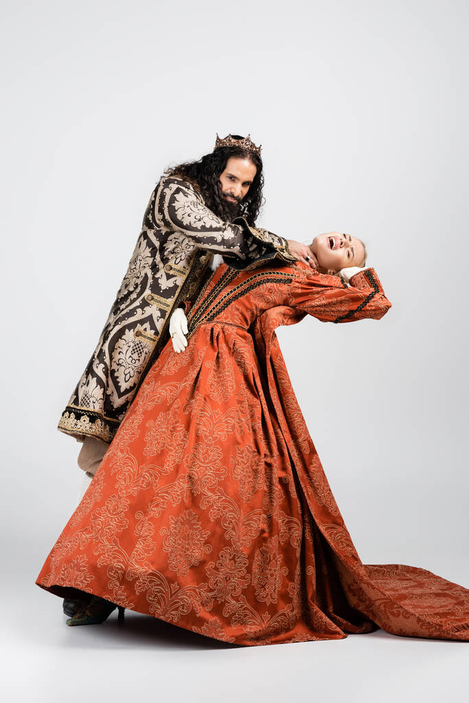 plná délka krutého hispánského krále ve středověkém oděvu udušení šokované královny ve zlaté koruně na bílém - Fotografie, Obrázek