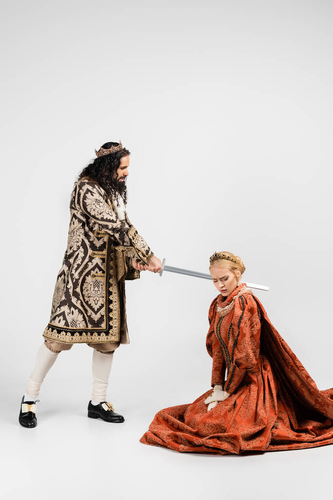 Orta çağ giysili zalim İspanyol kralın elinde beyaz taç giymiş korkmuş sarışın kraliçenin yanında. - Fotoğraf, Görsel