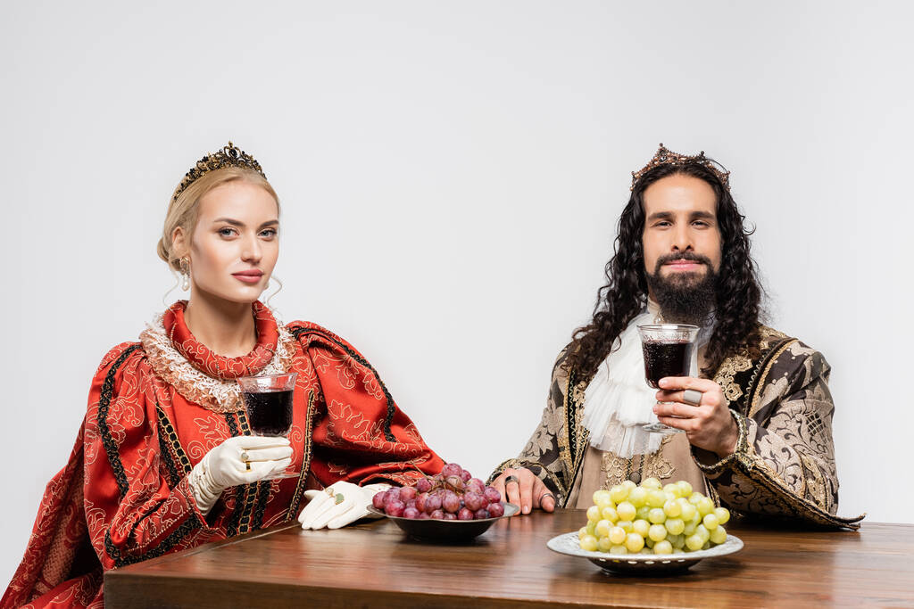 interracial couple historique en couronnes tenant des verres de vin rouge tout en étant assis à table avec des raisins isolés sur blanc - Photo, image