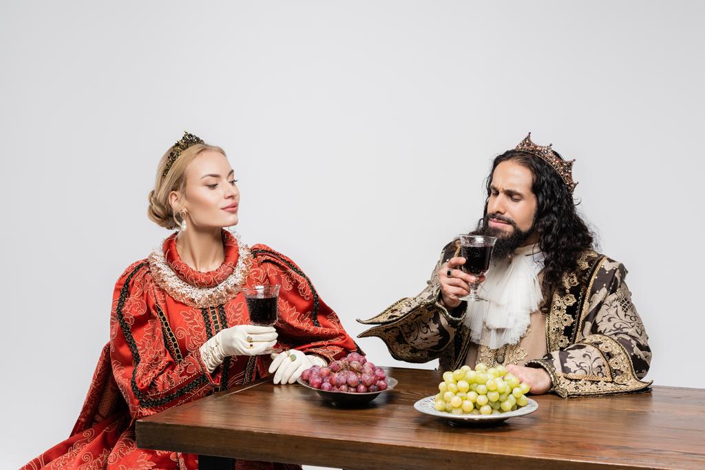 blond koningin kijken naar latino koning in middeleeuwse kleding ruiken rode wijn in glas geïsoleerd op wit - Foto, afbeelding