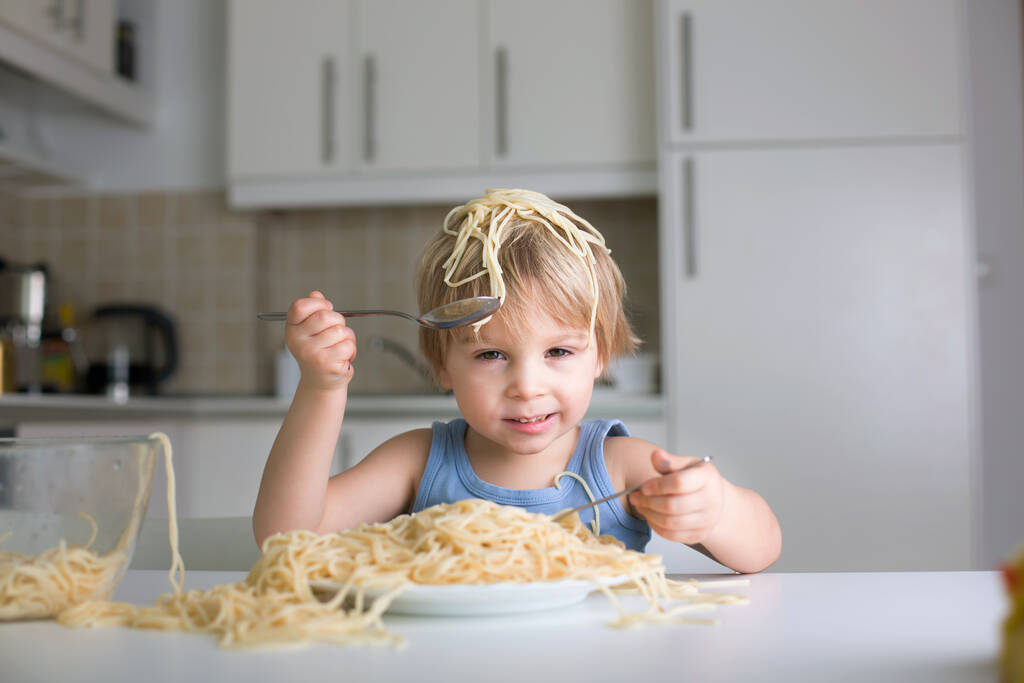 Kis szőke fiú, kisgyerek, spagettit eszik ebédre, és rendetlenséget csinál otthon a konyhában. - Fotó, kép