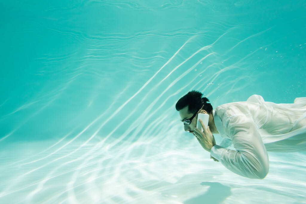 Muslim businessman in goggles talking on cellphone underwater in pool  - Photo, Image