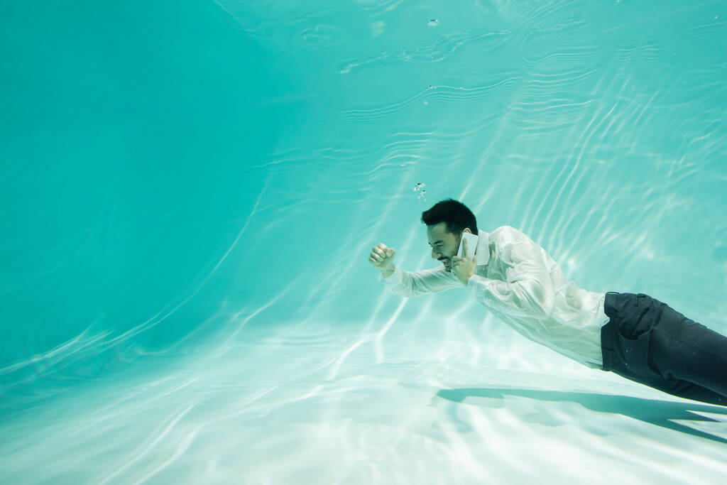 Alegre hombre de negocios árabe hablando por celular y mostrando sí gesto en la piscina  - Foto, imagen