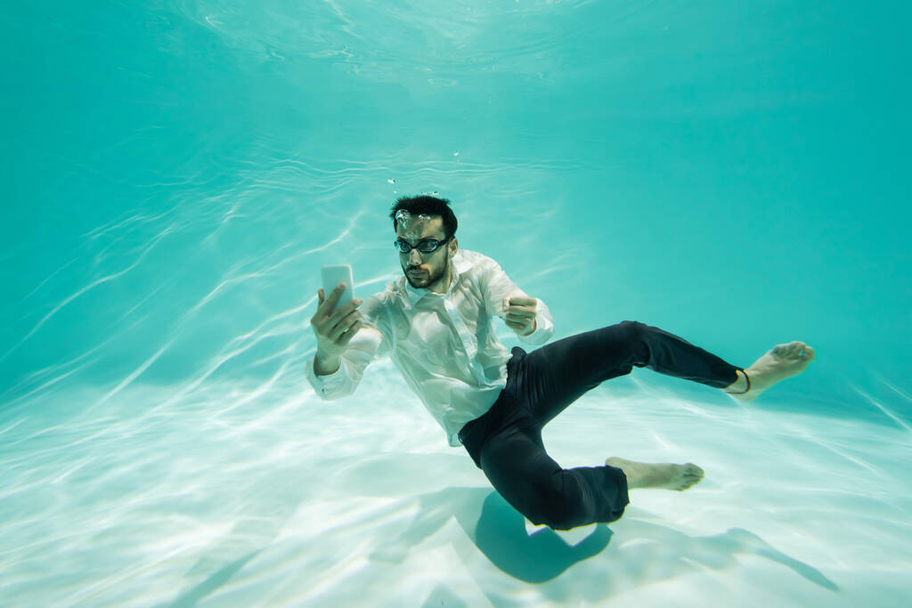 Arabian businessman in goggles looking at cellphone in swimming pool  - Photo, Image