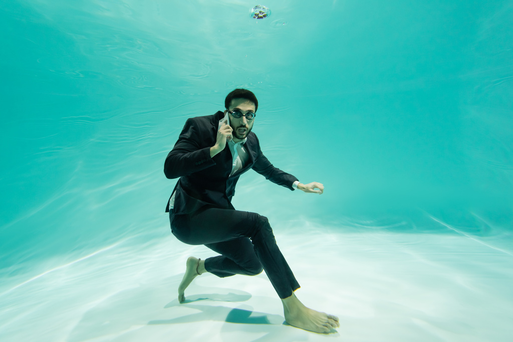 Arabische zakenman op zoek naar camera tijdens het praten op smartphone onder water in het zwembad  - Foto, afbeelding