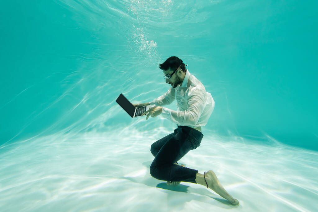 Μουσουλμάνος διευθυντής με επίσημη ένδυση και γυαλιά που χρησιμοποιεί φορητό υπολογιστή κάτω από το νερό  - Φωτογραφία, εικόνα