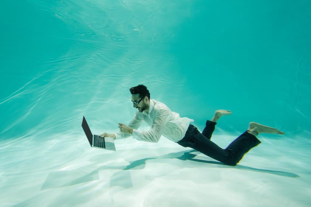 Hombre de negocios árabe sonriente sosteniendo portátil y nadando en la piscina  - Foto, imagen