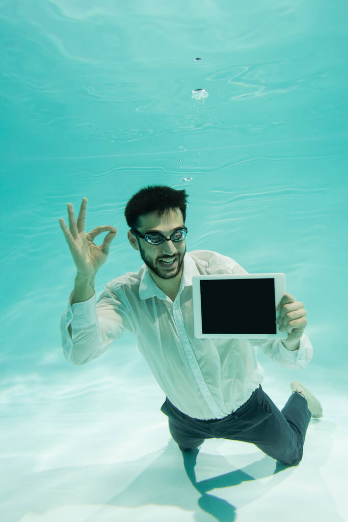Uomo d'affari musulmano sorridente mostrando gesto ok e tenendo tablet digitale sott'acqua  - Foto, immagini