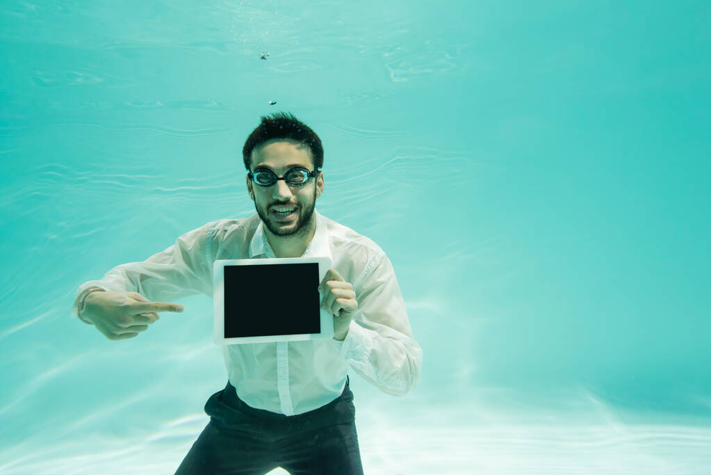 Arabischer Geschäftsmann lächelt und zeigt auf digitales Tablet mit leerem Bildschirm unter Wasser  - Foto, Bild