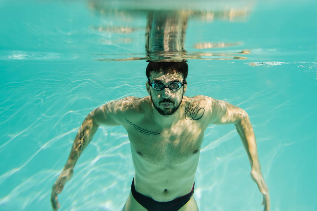Tätowierter arabischer Schwimmer mit Brille blickt in Kamera unter Wasser  - Foto, Bild