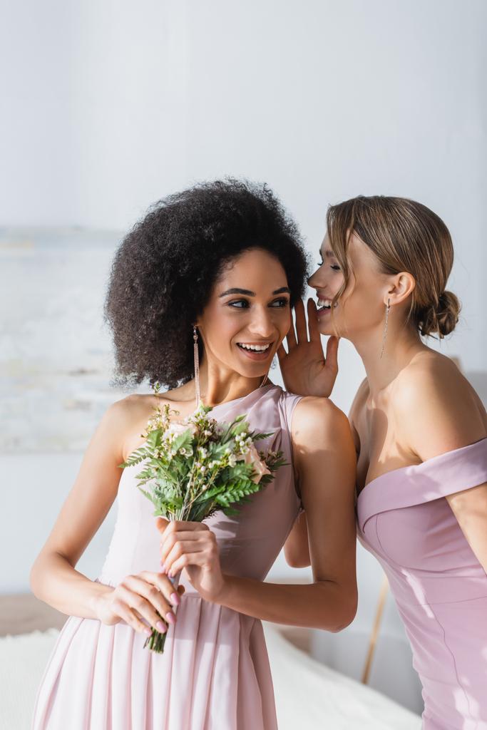 alegre mujer diciendo secreto a sonriente africano americano amigo celebración boda bouquet - Foto, Imagen