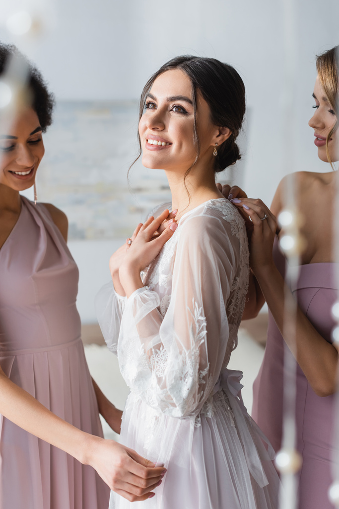 happy and dreamy bride looking away near smiling bridesmaids, blurred foreground - Photo, Image