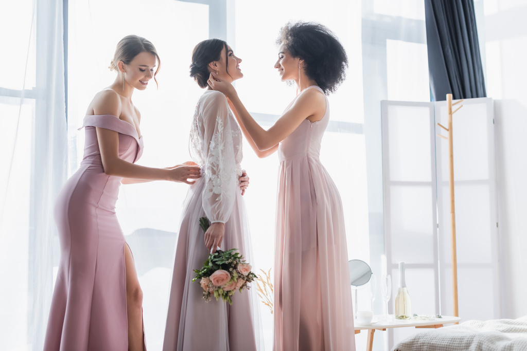 multicultural bridesmaids adjusting dress of happy bride holding wedding bouquet - Photo, Image