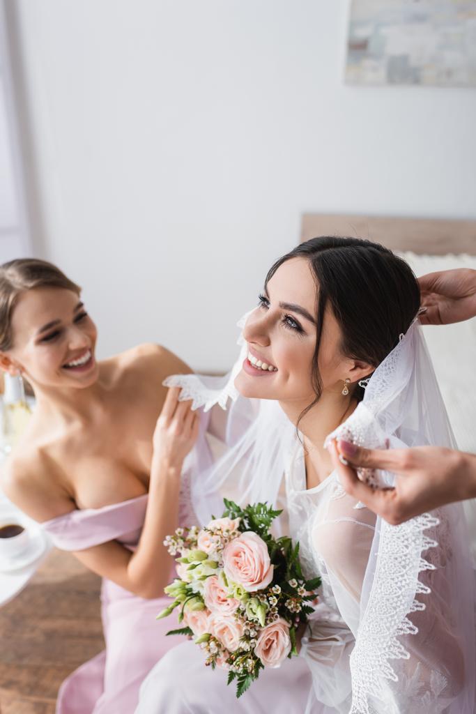 gelukkig bruid houden bruiloft boeket terwijl bruidsmeisjes repareren haar sluier - Foto, afbeelding