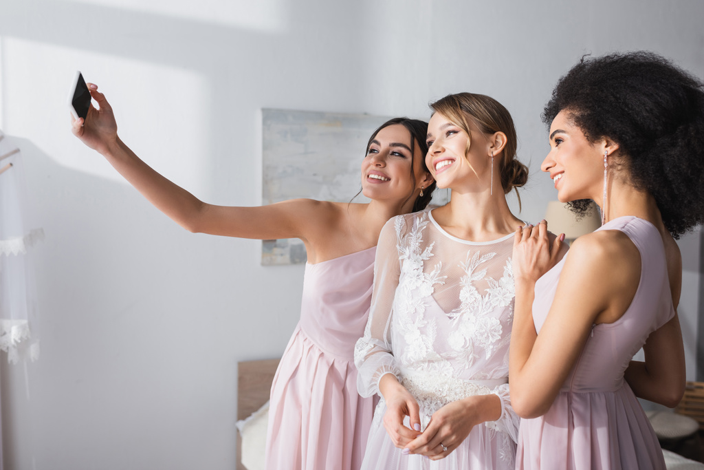 alegre mujer tomando selfie con novia y africano americano amigo en dormitorio - Foto, Imagen