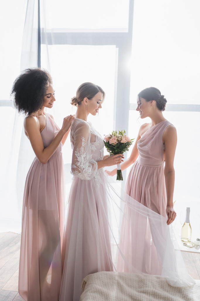 pretty bride holding wedding bouquet while interracial bridesmaids adjusting her dress on sunny morning - Photo, Image