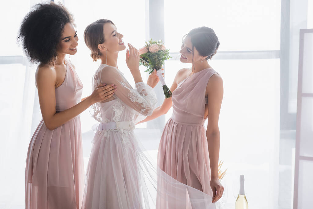 happy bride holding wedding bouquet and laughing with closed eyes near interracial bridesmaids - Photo, Image