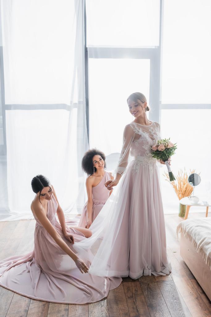 charming woman in wedding dress holding bouquet near bridesmaids at home - Photo, Image