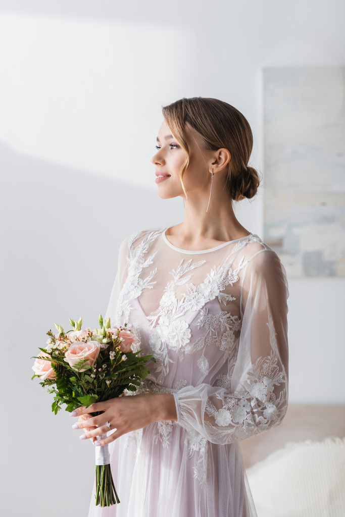 молода наречена в білій весільній сукні тримає букет і дивиться вдома
 - Фото, зображення