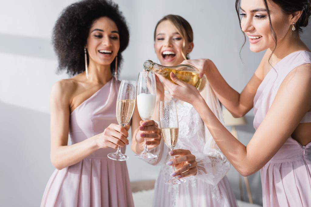 young woman pouring champagne near african american friend and bride on blurred background - Photo, Image