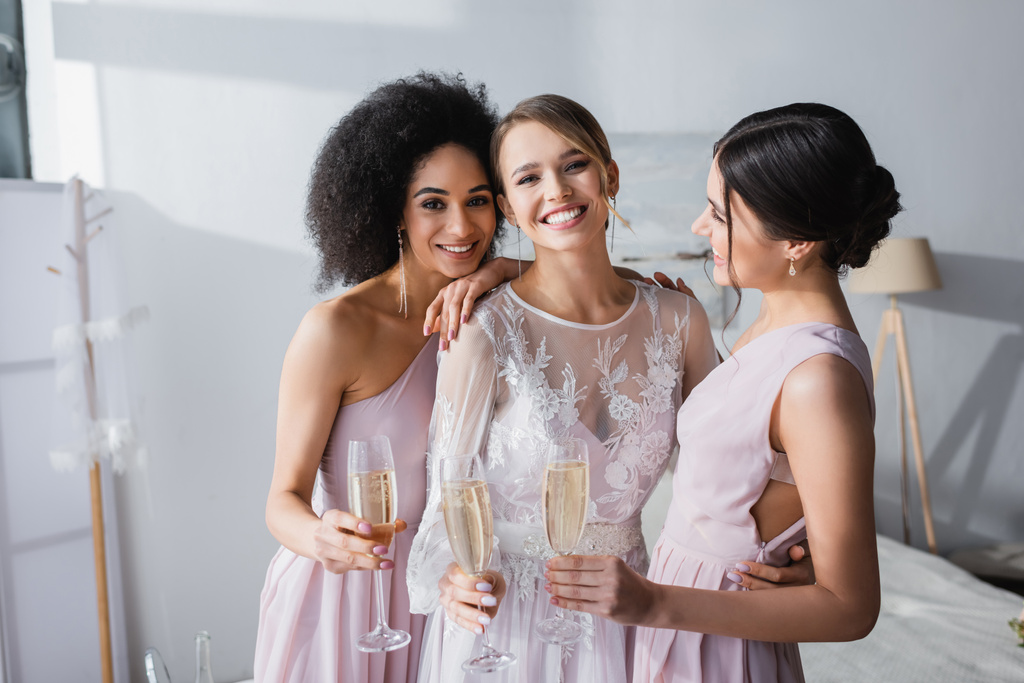 joyful bride looking at camera while holding champagne together with interracial friends - Photo, Image