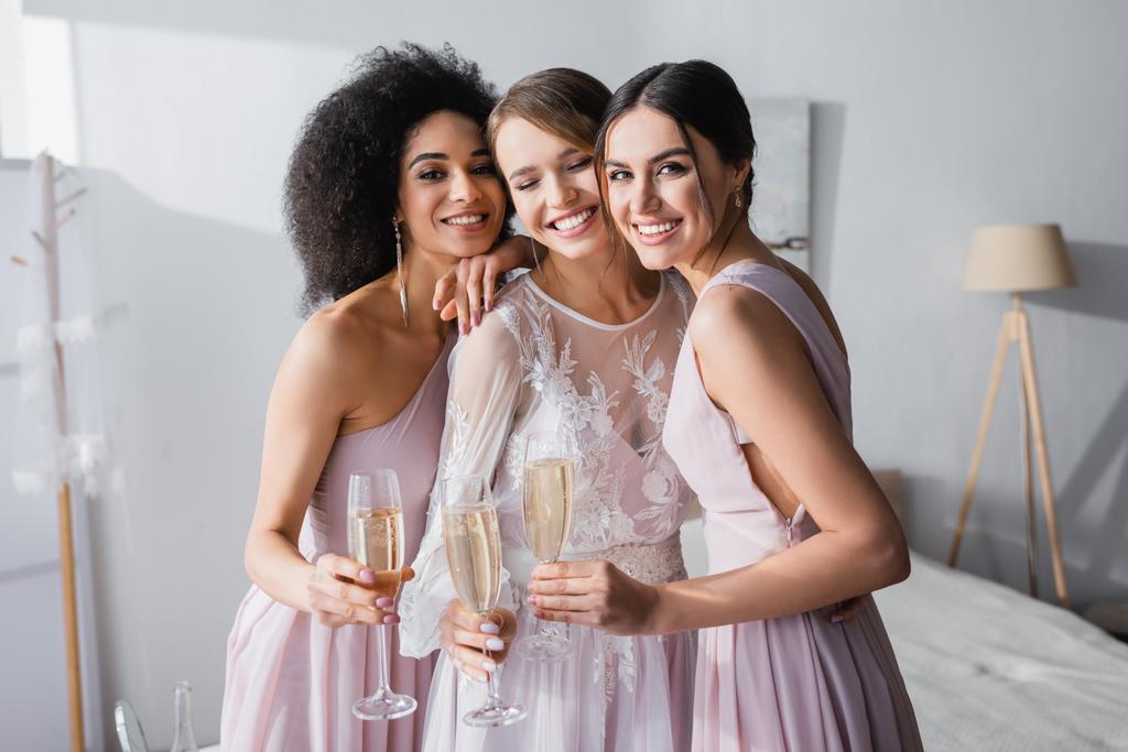 весёлые межрасовые подружки невесты, держащие бокалы шампанского возле счастливой невесты в спальне - Фото, изображение