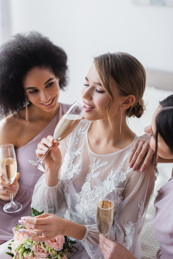 hezká nevěsta pije šampaňské v blízkosti multikulturní družičky v ložnici - Fotografie, Obrázek