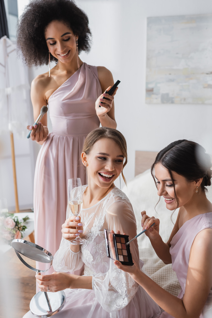 Lachende Braut hält Champagner und Spiegel in der Nähe interrassischer Freunde mit dekorativer Kosmetik - Foto, Bild