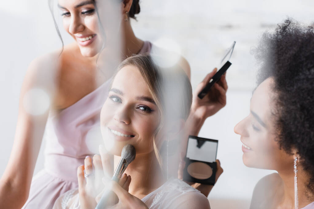 happy bride looking at camera while interracial women applying makeup, blurred foreground - Photo, Image