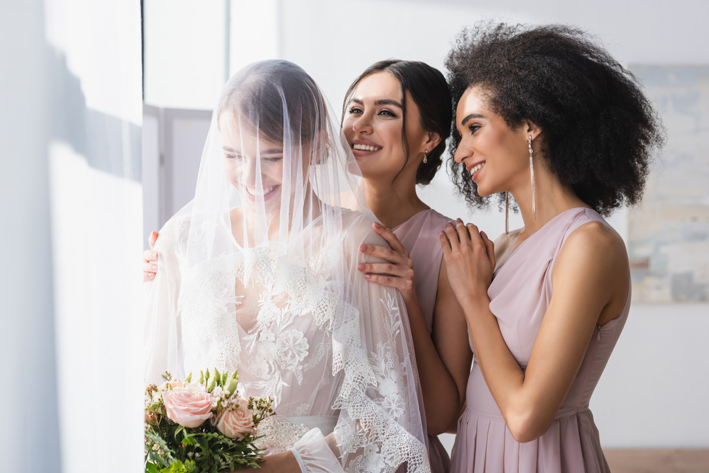 счастливая невеста, держащая свадебный букет возле улыбающихся межрасовых подружек невесты - Фото, изображение
