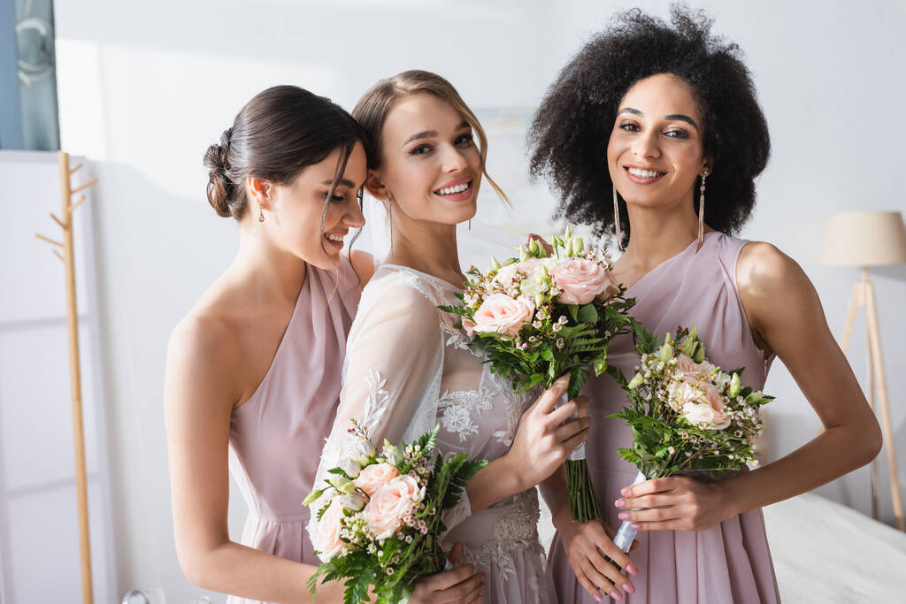pretty bride together with interracial bridesmaids holding wedding bouquets in bedroom - Photo, Image