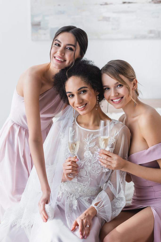 счастливые подружки невесты улыбаются в камеру, держа шампанское возле африканской американской невесты - Фото, изображение