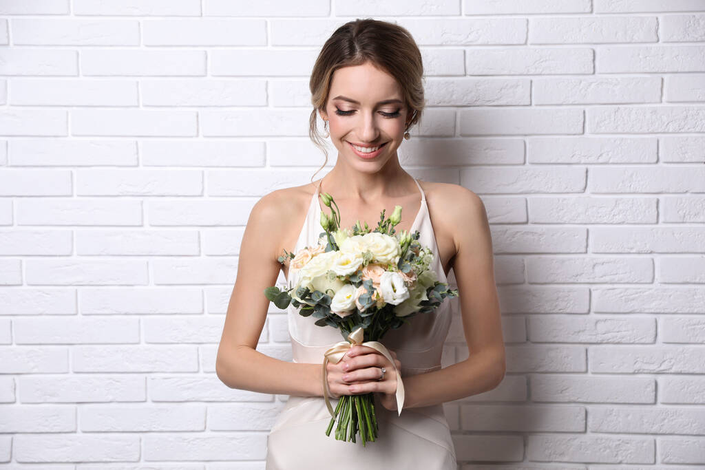 Mladá nevěsta s elegantním účesem drží svatební kytice v blízkosti bílé cihlové zdi - Fotografie, Obrázek