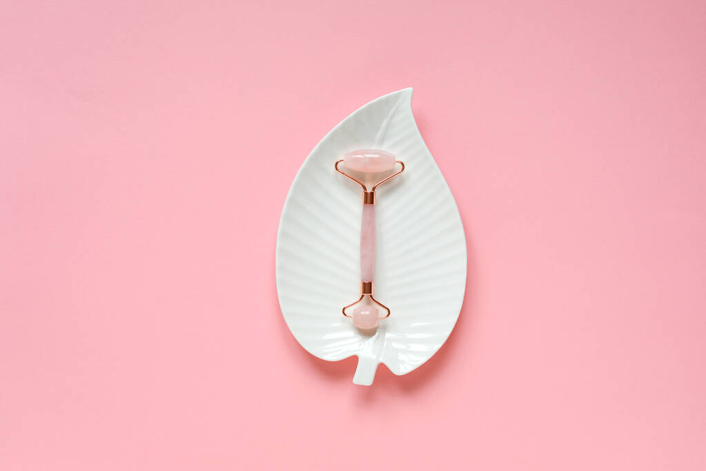 Rose quartz facial massage roller on white leaf shaped plate on pink background. Copy space. Beauty care accessory, skin massaging concept.  - Photo, Image