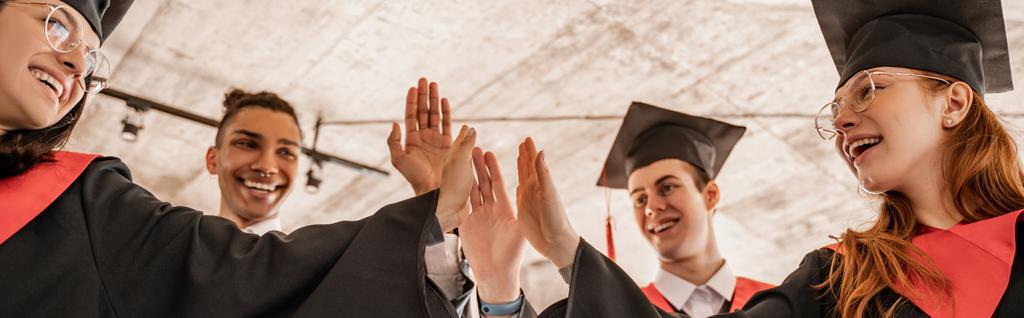 szczęśliwy międzyrasowy studenci w suknie i czapki posiadające dyplom i dając przybicie piątki, absolwent klasy 2021, sztandar - Zdjęcie, obraz