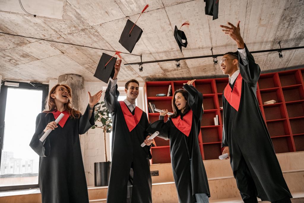 κατάπληκτοι διαφυλετικοί φοιτητές με φορέματα ρίχνουν καπέλα και κατέχουν δίπλωμα, τάξη αποφοίτησης 2021 - Φωτογραφία, εικόνα