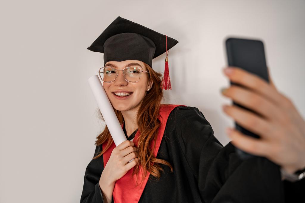 joyeux étudiant en chapeau de graduation et robe de prendre selfie, senior 2021 - Photo, image