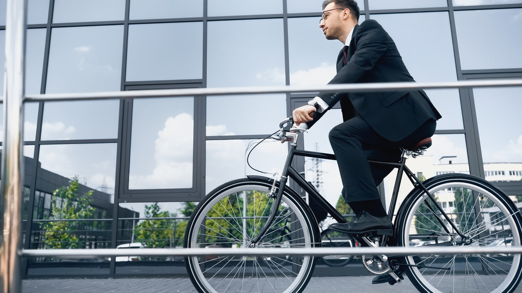 full length of businessman in suit and glasses riding bike near building with glass facade - Photo, Image