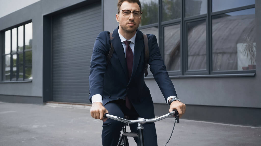 Geschäftsmann im Anzug blickt beim Fahrradfahren in der Nähe eines Gebäudes in die Kamera  - Foto, Bild