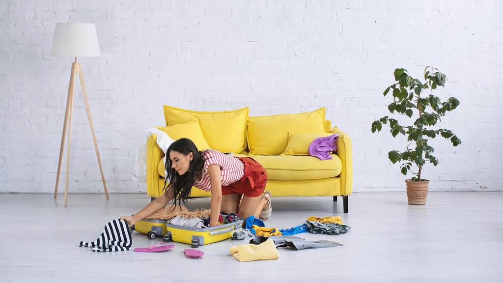 morena mujer embalaje equipaje con ropa de verano en el suelo en la sala de estar - Foto, imagen