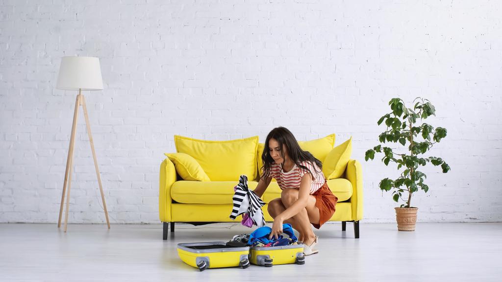 brunette woman packing yellow luggage near couch in living room  - Photo, Image