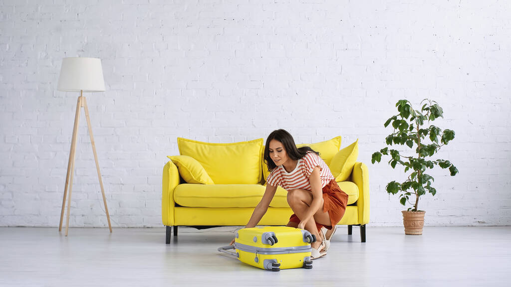 brunette woman smiling while looking at yellow luggage at home - Photo, Image