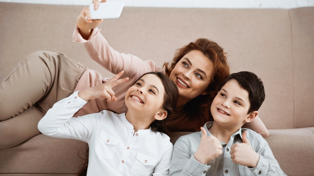 Wesołe dzieci gestykulują podczas robienia selfie z uśmiechniętą matką w domu  - Zdjęcie, obraz